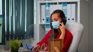 libero professionista Lavorando e parlando su Telefono seduta a posto di lavoro indossare protezione viso maschera durante coronavirus pandemia. donna chat con remoto squadra A proposito di su smartphone nel davanti di computer video