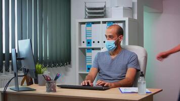 Coworkers with protection face masks working together in workplace during pandemic. Team in new normal office workspace in personal corporate company typing on computer keyboard looking at desktop video