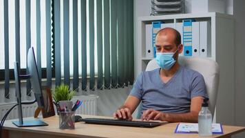 Businessman with medical protection face mask typing on computer in office room. Entrepreneur in new normal personal workplace corporate writing on pc keyboard looking at desktop video