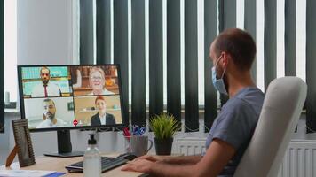 företag man med skyddande ansikte mask talande på ring upp på pc medan arbetssätt i ny vanligt kontor under coronavirus pandemisk. frilansare har uppkopplad konferens möte använder sig av internet teknologi video
