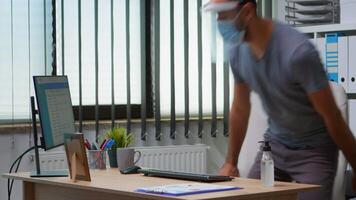 Man with protection mask and visor applying sanitizer gel rubbing hands before working at computer. Entrepreneur in new normal workplace disinfecting using antibacterial alcohol against corona virus. video