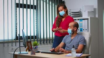 Workers talking wearing protection face masks in office room during coronavirus. Team in new normal office workspace in personal corporate company typing on computer keyboard and pointing at desktop video