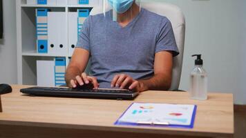 Busy employee wearing mask and visor in work place, typing and looking at camera. Entrepreneur in new normal personal corporate office writing on computer keyboard analyzing graphics video