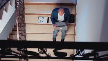superiore Visualizza di uomo d'affari seduta su le scale nel aziendale edificio analizzando documenti a partire dal appunti superlavoro nel finanziario azienda. ufficio esecutivo controllo annuale grafici in ritardo a notte. video
