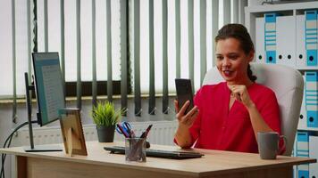 Hispanic woman having online meeting on phone sitting in modern office. Manager working with business remotely team discussing chatting having virtual conference, webinar using internet technology video