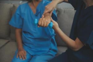 Old woman training with physiotherapist using dumbbells at home. Therapist assisting senior woman with exercises in nursing home. Elderly patient using dumbbells with outstretched arms. photo