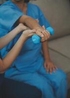 Old woman training with physiotherapist using dumbbells at home. Therapist assisting senior woman with exercises in nursing home. Elderly patient using dumbbells with outstretched arms. photo