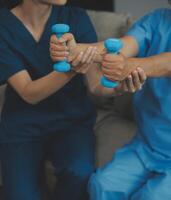 Old woman training with physiotherapist using dumbbells at home. Therapist assisting senior woman with exercises in nursing home. Elderly patient using dumbbells with outstretched arms. photo