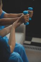 Old woman training with physiotherapist using dumbbells at home. Therapist assisting senior woman with exercises in nursing home. Elderly patient using dumbbells with outstretched arms. photo