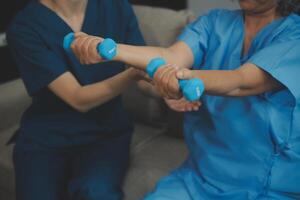 Old woman training with physiotherapist using dumbbells at home. Therapist assisting senior woman with exercises in nursing home. Elderly patient using dumbbells with outstretched arms. photo