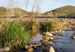 Scenic landscape of a small river with stones in the water, surrounded by green grass against the backdrop of beautiful hills under a cloudy sky photo