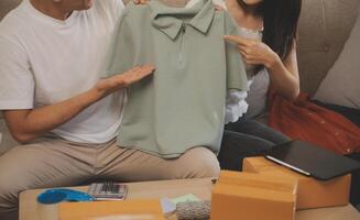 Two Asian blogger showing clothes in front of the camera to recording vlog live streaming at her shop. Online Shopping Cart notion. on sofa at home photo