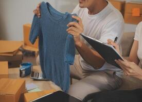 Two Asian blogger showing clothes in front of the camera to recording vlog live streaming at her shop. Online Shopping Cart notion. on sofa at home photo