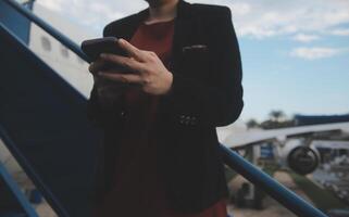 joven asiático mujer en internacional aeropuerto, utilizando móvil teléfono inteligente y comprobación vuelo a el vuelo información tablero foto