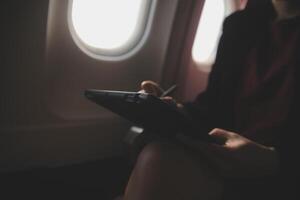 Blonde female tourist checking incoming notification on smartphone sitting on seat of airplane with netbook.Young businesswoman share media from telephone on laptop computer during plane flight photo