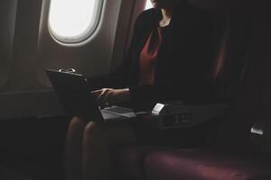 rubia hembra turista comprobación entrante notificación en teléfono inteligente sentado en asiento de avión con netbook.joven mujer de negocios compartir medios de comunicación desde teléfono en ordenador portátil computadora durante avión vuelo foto