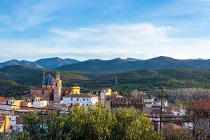 SONEXIA, VALENCIA, SPAIN - 12 april 2024 Beautiful picturesque landscape of a small town with a clock tower somewhere among the mountain hills covered with green trees photo