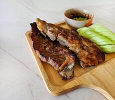 Grilled pork ribs, sliced cucumber and spicy sauce on wooden tray. Close up Barbecue food with fresh vegetables on white marble with copy space. photo