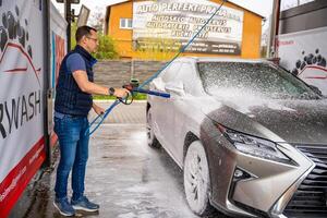 Prague, Czech Republic - April 5, 2024. Young man washes his car at a self-service car wash using a hose with pressurized water and foam. High quality photo