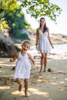 joven mujer madre con un pequeño hija en blanco vestidos teniendo un divertido en costa en el sombra de arboles y palmas foto