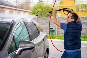 joven hombre lavados su coche a un autoservicio coche lavar utilizando un manguera con presurizado agua y espuma. foto