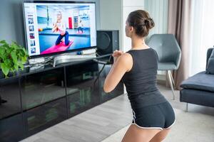 Young woman is doing fitness at home in living room while watching and participating in a class photo
