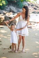 joven mujer madre con un pequeño hija en blanco vestidos teniendo un divertido en costa en el sombra de arboles y palmas foto