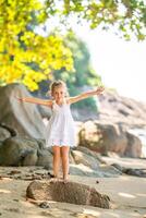 pequeño niña en blanco vestir teniendo divertido en costa en el sombra de arboles y palmas foto