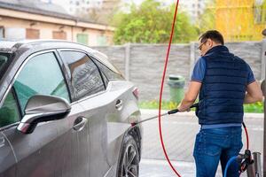 joven hombre lavados su coche a un autoservicio coche lavar utilizando un manguera con presurizado agua y espuma. alto calidad foto