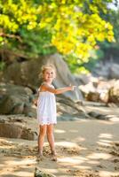 pequeño niña en blanco vestir teniendo divertido en costa en el sombra de arboles y palmas foto