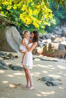 joven mujer madre con un pequeño hija en blanco vestidos en costa en el sombra de arboles y palmas alto calidad foto