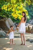 Young woman mother with a little daughter in white dresses walking on seashore in the shade of trees and palms photo