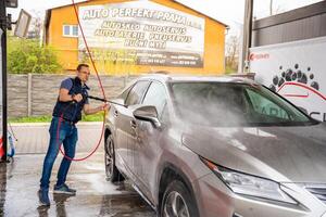 Prague, Czech Republic - April 5, 2024. Young man washes his car at a self-service car wash using a hose with pressurized water and foam. photo
