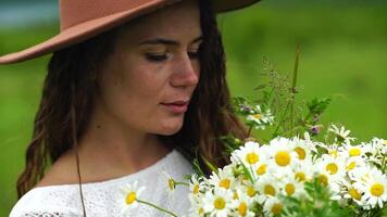 Chamomile woman. Happy woman in a white dress and brown hat holds a bouquet of daisies in her hands and smells them video