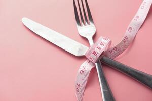 Knife and fork wrapped in tape measure on pink background. photo
