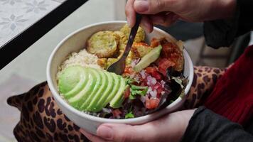 A young woman enjoys her dinner in a beautiful cafe where poke bowls are served. Hand is getting a piece of the poke bowl with fork. Hawaiian meal with falafel, rice, avocado, tofu, vegetables video