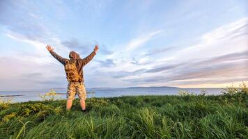 Man hiker in camo shorts and backpack on top of green hill, hands up, view on wild Atlantic way at Galway, Ireland, freedom, adventure and lifestyle concept, nature background photo