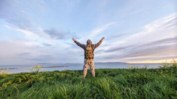 Man hiker in camo shorts and backpack on top of green hill, hands up, view on wild Atlantic way at Galway, Ireland, freedom, adventure and lifestyle concept, nature background photo