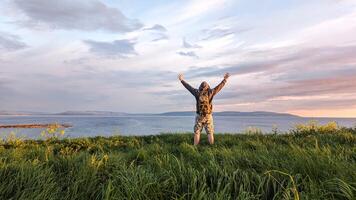 hombre caminante en camuflaje pantalones cortos y mochila en parte superior de verde colina, manos arriba, ver en salvaje atlántico camino a galway, Irlanda, libertad, aventuras y estilo de vida concepto, naturaleza antecedentes foto