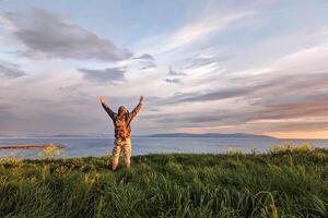 Man hiker in camo shorts and backpack on top of green hill, hands up, view on wild Atlantic way at Galway, Ireland, freedom, adventure and lifestyle concept, nature background photo