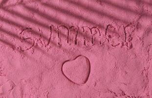 The word Summer handwritten and heart shape on pink sand surface with tropical palm tree leaf shadow. Minimal concept photo of summer vacation. Sun and shadows. Exotic flat lay. Summer love aesthetic.