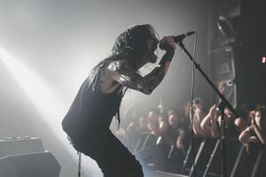 A rock frontman singing to the microphone to the crowd on a concert in a stage backlights. Post hardcore style. Back view photo