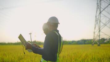 Two men are standing in a field, one of them is holding a piece of paper. They are wearing safety gear and seem to be discussing something video