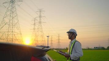 A man in a safety vest is standing next to a car and looking at a tablet. The scene is set in a field with power lines in the background. Scene is serious and focused video