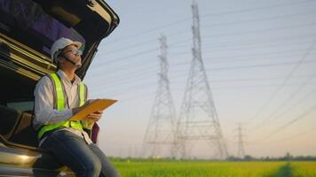 A man in a safety vest is sitting in a car and writing on a clipboard. The scene is set in a field with tall power lines in the background. Scene is serious and focused video