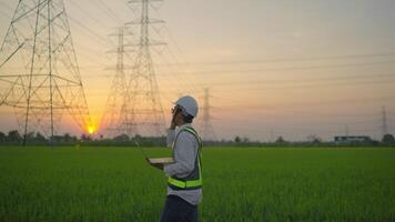 A man wearing a safety vest is standing in a field with a laptop in his hand. The sky is orange and the sun is setting. The man is likely working on a project related to the field video