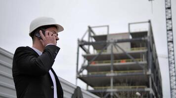 un arquitecto en un negocio traje y un blanco casco, participación un teléfono en su mano y hablando a alguien, en el antecedentes en frente él es líder el construcción de edificios y un construcción grua. video