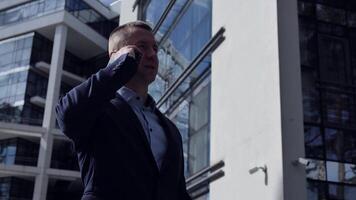 Businessman swiftly raises phone to ear backdrop of office building. Businessman engages in serious dialogue, intensity marked. Businessmans strategic conversation underscores his focus and expertise. video