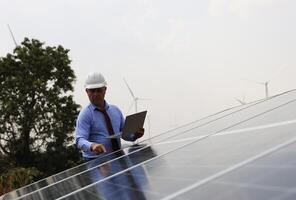 A senior male engineer inspects solar panels on the windmill farm. Clean energy. Engineers inspect a solar panel system with a turbine farm in the background, a renewable energy concept photo
