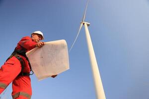 windmill engineer with red safety uniform hold drawing work at front of wind turbine farm photo